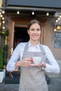 Smiling cute waitress in grey apron offering cup of coffee