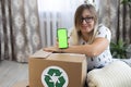 Smiling cute woman holds a phone with a chromakey screen in her hand over a box with a recycling sign. Clothes are Royalty Free Stock Photo