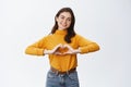 Smiling cute woman showing heart sign, I love you gesture, staring at camera with happy face, standing against white Royalty Free Stock Photo