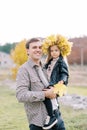 Smiling dad holding in his arms a little girl in a wreath of autumn leaves with a yellow leaf in her hand