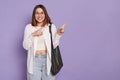 Smiling delighted beautiful woman with black bag wearing white shirt and jeans posing isolated over purple background, pointing Royalty Free Stock Photo