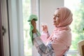 Smiling cheerful Arab Muslim woman with covered head in hijab washing and wiping windows in the veranda during spring cleaning