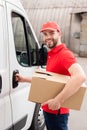 smiling delivery man in uniform with cardboard box standing near white van Royalty Free Stock Photo