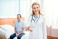 Smiling doctor with stethoscope standing in chamber in hospital, with patient behind Royalty Free Stock Photo