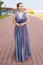 Smiling dreamy young Caucasian woman in amethystine floor-length sparkly dress on pavement