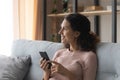 Smiling dreamy Latin woman sit on sofa holding modern cellphone
