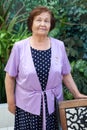 Smiling elder woman standing next to chair, portrait in greenhouse, looking at camera