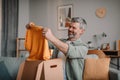 Smiling elderly european man with beard unpacks cardboard box with clothes in room interior