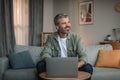 Smiling elderly european man with beard in wireless headphones typing on laptop and looking at empty space Royalty Free Stock Photo