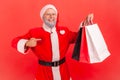 Smiling elderly gray bearded man in santa claus costume pointing finger at paper shopping bags in his hand, doing holidays