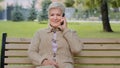 Smiling elderly gray-haired lady with beautiful face sitting on park bench takes phone call, woman of retirement age Royalty Free Stock Photo