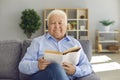 Smiling elderly man or grandfather in blue shirt sitting on sofa and reading book at home Royalty Free Stock Photo