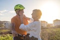 Smiling elderly woman takes care of her cyclist husband by putting on his helmet before riding his bike. Bright sunset light