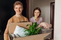 Smiling energetic students teenagers man, woman hold carton box with flower and clothes. Changeover new university room