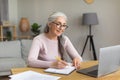 Smiling european old woman with gray hair in glasses look at computer, make notes at table Royalty Free Stock Photo