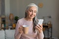 Smiling european old woman with gray hair looks at smartphone, have chat and drinks coffee in room interior