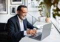 Smiling european senior businessman in suit typing on laptop at table in modern office interior Royalty Free Stock Photo