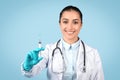 Smiling european woman doctor wearing sterile gloves, holding medical syringe, blue studio background Royalty Free Stock Photo