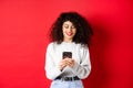 Smiling european woman reading mobile phone screen, texting a message on smartphone, standing in sweatshirt against red Royalty Free Stock Photo