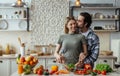Smiling european young guy with stubble hugs, kisses blonde female, lady preparing salad at table