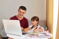 Smiling father and a little daughter doing homework at home, sitting at table in room, looking at laptop screen with smiles, Royalty Free Stock Photo