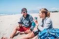 Smiling Father in sunglasses with smartphone sitting with teenager son with backpacks on the sandy seaside beach. Boy drinking wat Royalty Free Stock Photo