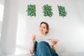 Smiling female artist sitting on bed with paintbrush, palette under her hand-drawn painting art works on the wall at