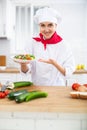 Smiling female chef in white uniform offering plate of vegetable salad in home kitche