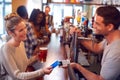 Smiling Female Customer In Bar Making Contactless Payment With Card For Drinks To Male Bartender