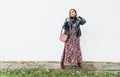 Smiling female dressed boho fashion style colorful long dress with black leather biker jacket with brown leather flap bag posing
