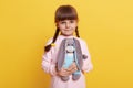 Smiling female kid with pigtails wearing pale pig shirt posing isolated over yellow background, holding soft rabbit toy in hands