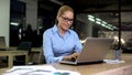 Smiling female working on project in office, using laptop at nigh shift, job