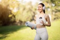 Smiling fit woman with headphones and a yoga mat enjoys a sunny day in the park, ready for a wellness workout Royalty Free Stock Photo
