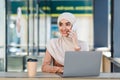 Smiling friendly cute millennial arab muslim woman in hijab speaks on phone and types on laptop in cafe