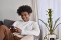 Smiling gen z African American teen sitting on couch using smartphone. Royalty Free Stock Photo