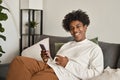 Smiling gen z African American teen sitting on couch using smartphone. Royalty Free Stock Photo