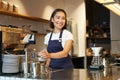 Smiling girl barista in cafe, preparing cappuccino in coffee machine, steaming milk, wearing uniform apron Royalty Free Stock Photo