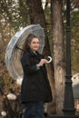 Smiling girl in black coat peeks out from under transparent umbrella. Portrait of young woman against yellow trees Royalty Free Stock Photo