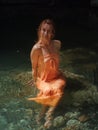 Smiling girl in orange sundress sits in clear river water in rocky lagoon Royalty Free Stock Photo