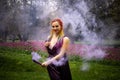 A girl with a smile with bright makeup and colored braids in a purple dress blows artificial purple smoke in a spring Royalty Free Stock Photo