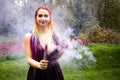 A girl with a smile with bright makeup and colored braids in a purple dress blows artificial purple smoke in a spring Royalty Free Stock Photo