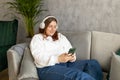 Smiling girl relaxing at home, she is listening to music, using a smartphone and wearing white headphones. Young 30s Royalty Free Stock Photo