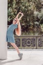 A smiling girl in a short dress is posing leaning on a column standing on her toes and raising her hands up against the