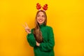 Smiling girl shows peace v-sign gesture, wears sweater, Christmas reindeer antlers and looks up on yellow background. Royalty Free Stock Photo