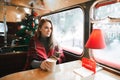 Smiling girl sitting with cup of coffee in atmospheric cafe with Christmas tree, holding smartphone and looking out the window