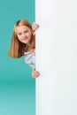 A smiling girl stands behind a white blank panel isolated against a turquoise background. Looks out from behind the banner, an Royalty Free Stock Photo