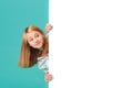 A smiling girl stands behind a white blank panel isolated against a turquoise background. Looks out from behind the banner, an Royalty Free Stock Photo