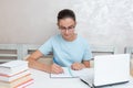 A smiling girl student sitting at a table with a laptop writes a task in a notebook. Girl student studies at home. Royalty Free Stock Photo