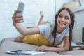 smiling girl taking selfie on smartphone while lying on sofa with copybook