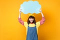 Smiling girl teenager in french beret, denim sundress hold blue empty blank Say cloud, speech bubble isolated on yellow Royalty Free Stock Photo